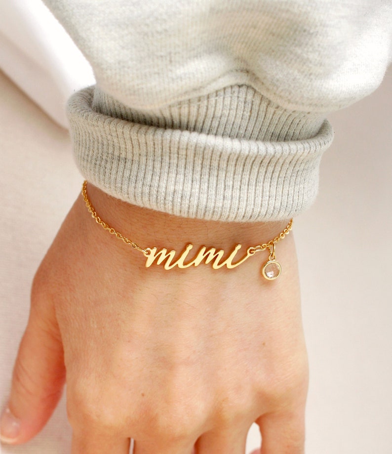 Op de afbeelding: Gouden ketting armband met de naam "mimi" in cursief schrift. Een kleine heldere kristallen hanger hangt aan de ketting.