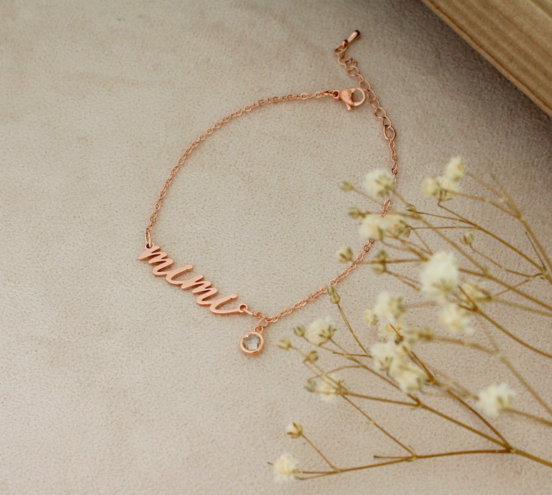 Op de afbeelding: Een ros&eacute;gouden armband met het woord "mimi" in cursief schrift en een kleine, heldere kristallen bedel.