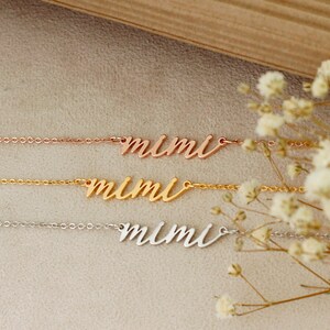 Op de afbeelding: Drie delicate kettingen met de naam "mimi" in cursief schrift. De kettingen liggen op elkaar op een lichtbruin oppervlak met witte bloemen op de achtergrond. De bovenste ketting is ros&eacute;goud, de middelste ketting is goud en de onderste ketting is zilver.