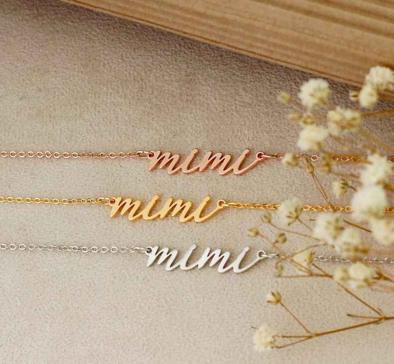Op de afbeelding: Drie delicate kettingen met de naam "mimi" in cursief schrift. De kettingen liggen op elkaar op een lichtbruin oppervlak met witte bloemen op de achtergrond. De bovenste ketting is ros&eacute;goud, de middelste ketting is goud en de onderste ketting is zilver.