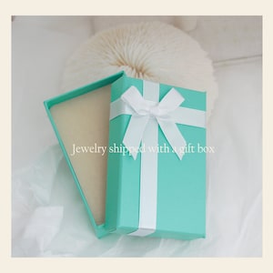 Puede incluir: Una caja de regalo turquesa con una cinta y un lazo blancos. La caja est&aacute; abierta y el interior est&aacute; forrado con papel de seda blanco. "Jewelry shipped with a gift box" est&aacute; escrito en el papel de seda.