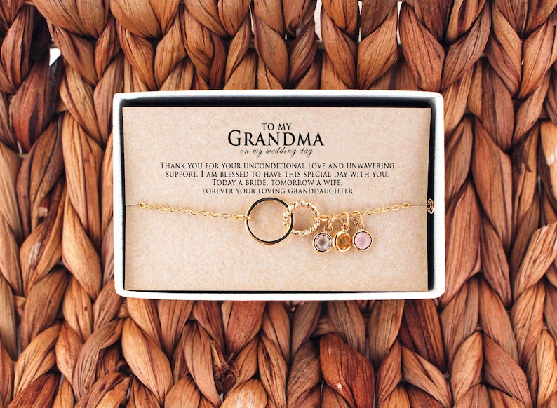 Puede incluir: Una caja blanca con un brazalete dorado en su interior. El brazalete tiene un dije circular, un dije retorcido y tres dijes peque&ntilde;os con piedras preciosas. La caja contiene una tarjeta con el texto "A mi abuela en mi d&iacute;a de boda. Gracias por tu amor incondicional y tu apoyo inquebrantable. Soy bendecida de tener este d&iacute;a especial contigo. Hoy una novia, ma&ntilde;ana una esposa. Por siempre tu nieta cari&ntilde;osa."