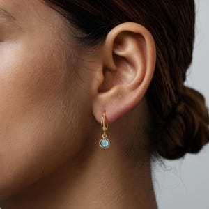 Pode incluir: Brincos de argola de ouro com uma pequena gema azul claro pendurada na parte inferior.