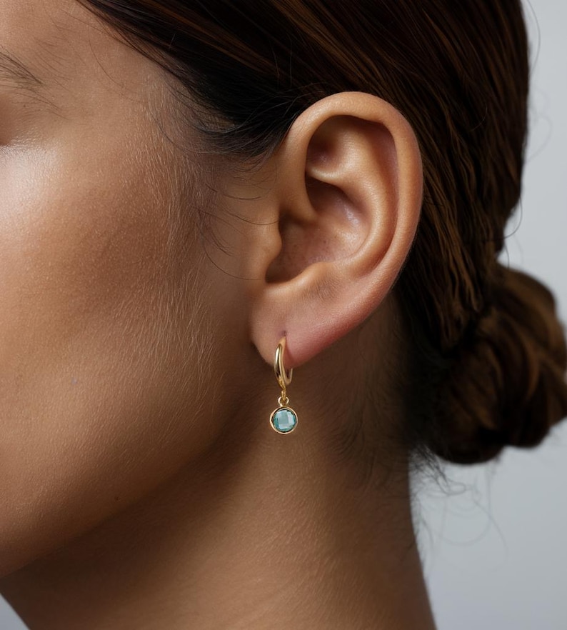 Pode incluir: Brincos de argola de ouro com uma pequena gema azul claro pendurada na parte inferior.