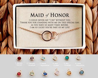 May include: A gold necklace with two connected circles hangs on a white card with the text "Maid of Honor" and a message thanking the recipient for being a part of the special day. Below the card are twelve small charms with different colored gemstones, each labeled with a month and gemstone name.