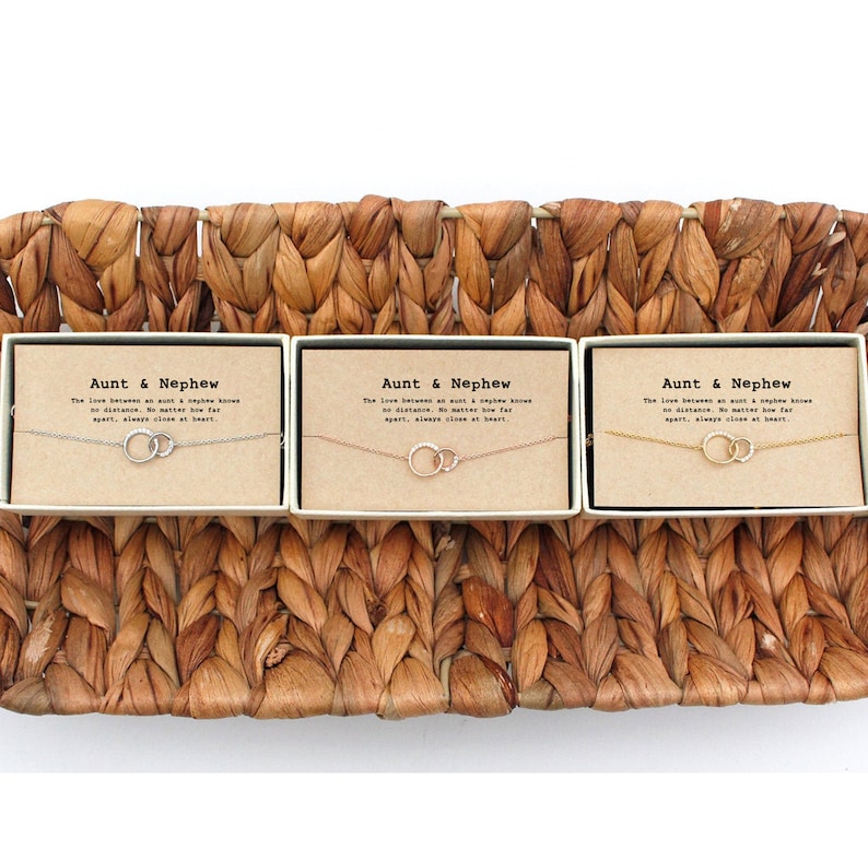 May include: Three small gift boxes with brown paper wrapping and white text that reads "Aunt & Nephew" and a quote about the love between an aunt and nephew. Each box contains a delicate silver or gold chain necklace with two interlocking circles.