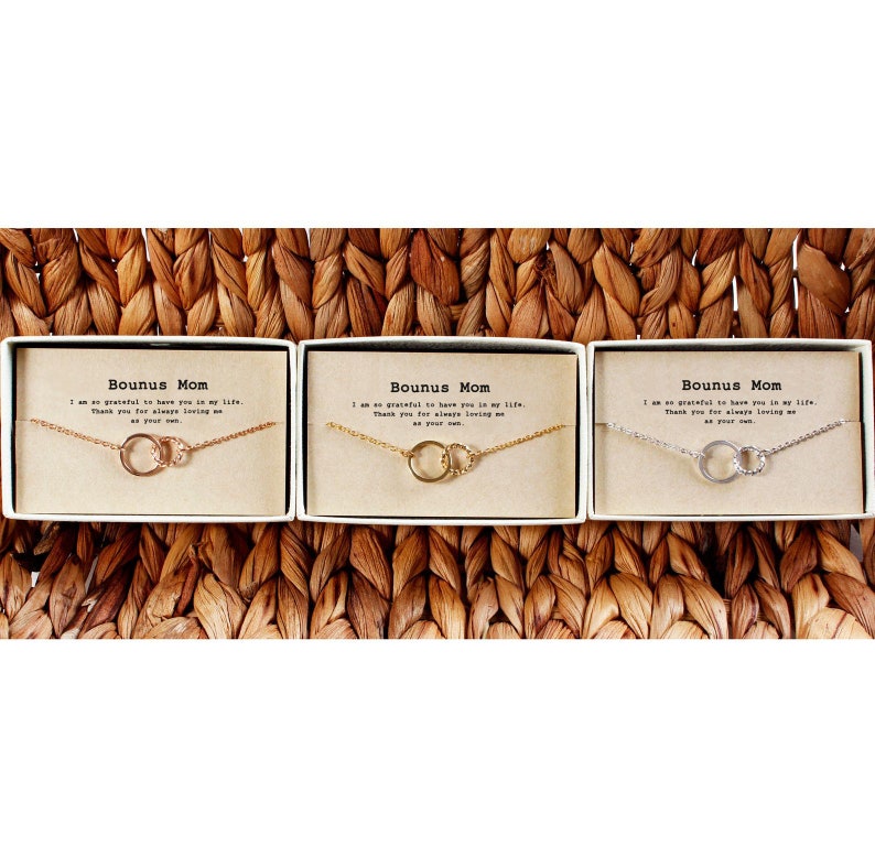 May include: Three small gift boxes with the text "Bounus Mom" on the front. Each box contains a delicate necklace with two interlocking circles. The necklaces are available in gold, silver, and rose gold.