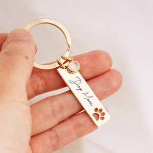 May include: Gold-tone keychain with a rectangular charm that says "Dog Mom" with a paw print cutout. The charm has a small clear crystal attached to it.