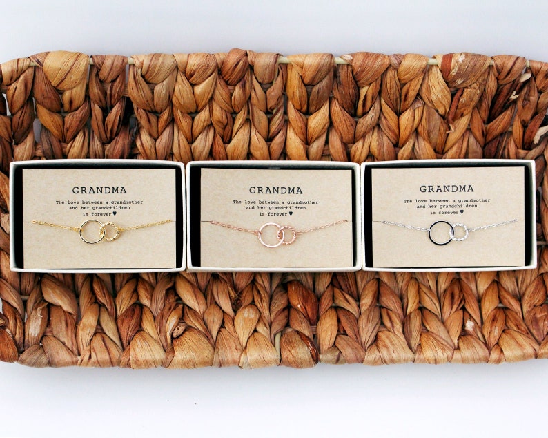 May include: Three small gift boxes with the word "Grandma" printed on the front. Each box contains a delicate silver chain necklace with two connected circles. The necklaces are in gold, rose gold, and silver.