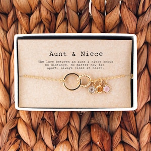 May include: A gold chain bracelet with a charm that says "Aunt & Niece" and features a circle charm with three small gemstone charms. The bracelet is in a small box with a card that reads "The love between an aunt & niece knows no distance. No matter how far apart, always close at heart."