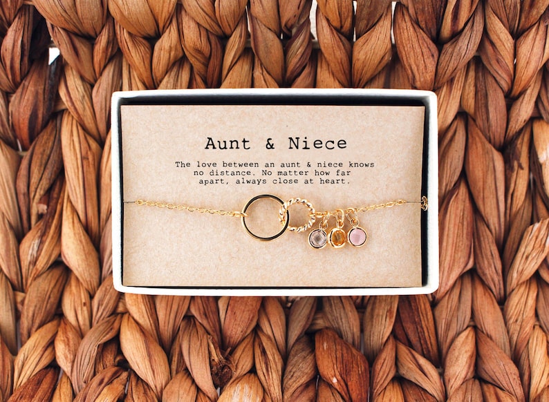 May include: A gold chain bracelet with a charm that says "Aunt & Niece" and features a circle charm with three small gemstone charms. The bracelet is in a small box with a card that reads "The love between an aunt & niece knows no distance. No matter how far apart, always close at heart."