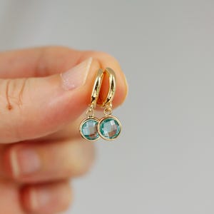 Pode incluir: Brincos de argola de ouro com uma gema azul pendurada. Os brincos s&atilde;o pequenos e delicados.