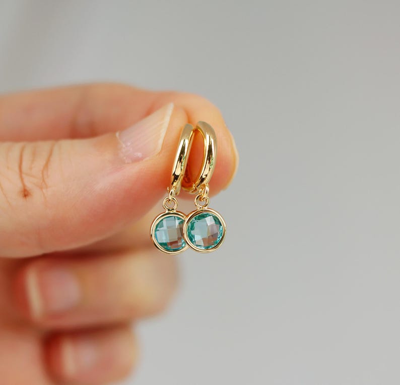Pode incluir: Brincos de argola de ouro com uma gema azul pendurada. Os brincos s&atilde;o pequenos e delicados.