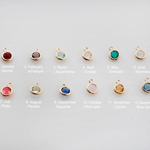 May include: Twelve gold charms with different colored gemstones, each representing a different month of the year. The charms are arranged in two rows of six. The first row includes January (garnet), February (amethyst), March (aquamarine), April (crystal), May (emerald), and June (moonstone). The second row includes July (ruby), August (peridot), September (sapphire), October (pink opal), November (citrine), and December (blue topaz).