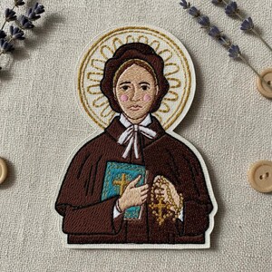 May include: An embroidered patch depicting a figure in a brown habit with a white collar and bow, holding a blue book with a gold cross and a gold rosary. The figure has a gold halo behind their head and pink cheeks on a white fabric background.