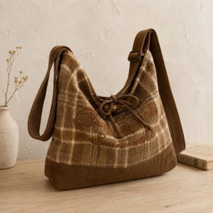 May include: A brown and beige plaid hobo bag featuring a corduroy base and matching shoulder strap. The bag has a drawstring closure with ties at the center and embroidered text that reads Best starts the sunrise with.