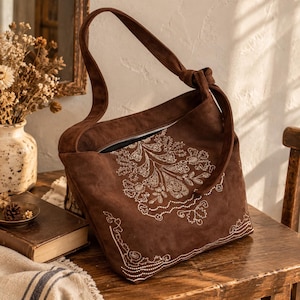 Könnte beinhalten: Eine dunkelbraune Hobo-Tasche aus Wildleder mit einem einzelnen geknoteten Schulterriemen und einem Reißverschluss oben. Die Vorderseite der Tasche ist mit komplizierten weißen Blumenstickereien verziert, die sich über die Mitte und die unteren Ecken erstrecken.