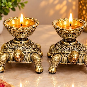 May include: Two gold-toned metal turtle-shaped oil lamps, or diyas, featuring intricate embossed patterns on the shells and legs. Each turtle supports a wide, decorative bowl at the top, holding a lit flame fueled by oil, creating a warm glow.