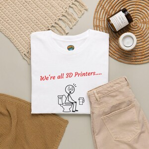 May include: A white t-shirt featuring the red text "We're all 3D Printers...." above a black line drawing of a stick figure sitting on a toilet with a roll of toilet paper nearby. The shirt has a small circular logo on the inside neck label.