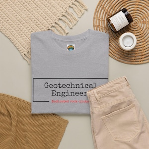 Może przedstawiać: Szary melanżowy t-shirt z czarnym prostokątnym obrysem zawierającym czarny napis Geotechnical Engineer oraz czerwony napis Dedicated rock-licker poniżej. Koszulka posiada małe okrągłe logo na wewnętrznej metce przy kołnierzu.
