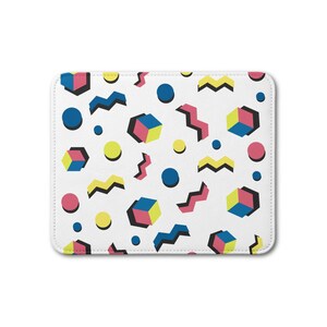 Könnte beinhalten: Ein rechteckiges Mauspad mit weißem Hintergrund und einem sich wiederholenden Muster aus bunten geometrischen Formen, darunter blaue, gelbe und rosa Kreise, gelbe und blaue Würfel sowie zickzackförmige Linien in Pink, Blau und Gelb.