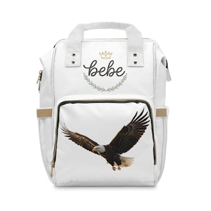 Könnte beinhalten: Ein weißer Rucksack mit einem Tragegriff und einer Vordertasche. Der obere Bereich zeigt ein goldenes Kronensymbol über dem Wort "bebe" in schwarzer Schreibschrift, umrahmt von einem Lorbeerkranz. Die Vordertasche zeigt eine Grafik eines Weißkopfseeadlers.