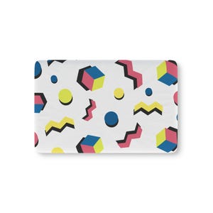 Könnte beinhalten: Eine rechteckige Matte mit weißem Hintergrund und einem wiederkehrenden Muster aus geometrischen Formen, darunter blaue und rosa Würfel, gelbe Kreise sowie gezackte Zickzacklinien in Pink und Gelb, die mit kräftigen schwarzen Schatten versehen sind.