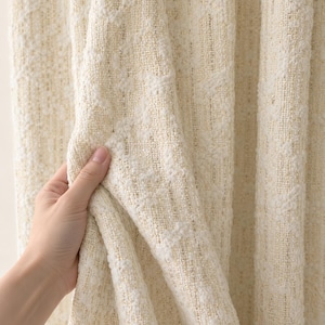 May include: A cream-colored fabric curtain featuring a textured, woven surface with a subtle, repeating pattern. The material has a thick, tactile quality with visible interlaced threads and a soft, off-white hue throughout the entire drape.