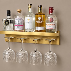 Puede incluir: Un estante de pared de metal dorado con una superficie superior que sostiene cinco botellas de ginebra y vodka, y cuatro copas de cristal transparente con patrón de diamante que cuelgan boca abajo del soporte integrado.
