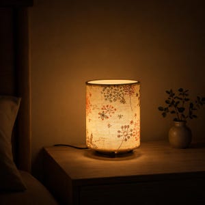 Könnte beinhalten: Eine zylindrische Tischleuchte mit einem hellen Lampenschirm, der mit einem zarten Muster aus orangefarbenen und dunkelbraunen löwenzahnartigen Blumenmotiven verziert ist. Die Lampe strahlt ein warmes, sanftes Licht in die Umgebung ab.