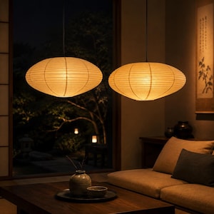 May include: Two oval-shaped pendant lights with ribbed paper shades emit a warm, soft glow. The fixtures feature a horizontal structure with thin, visible wire ribbing, suspended from the ceiling to provide ambient lighting in a room.