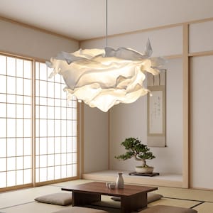 Op de afbeelding: Een grote, witte, sculpturale hanglamp met meerdere gelaagde, vloeiende, stofachtige panelen die een zachte, diffuse gloed creëren. De lamp hangt aan het plafond aan een dunne, rechte, metalen stang in een centrale positie.
