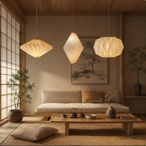 Op de afbeelding: Drie gebroken witte hanglampen met geplooide papieren kappen hangen aan het plafond. De linkerlamp is ruitvormig, de middelste lamp is langwerpig ovaal en de rechterlamp is een brede, afgeronde bol met verticale plooien.