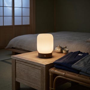 May include: A cylindrical table lamp with a ribbed, frosted white glass shade and a dark brown wooden base. The lamp emits a warm, soft glow and features a small circular power button on the front of the wooden base, resting on a wooden table.