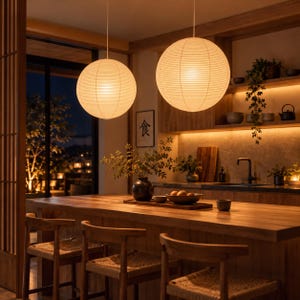Op de afbeelding: Twee grote, bolvormige papieren hanglampen met een geribbelde textuur hangen boven een houten kookeiland. De warm gloeiende lampen verlichten een keukenruimte met houten kasten, barkrukken met gevlochten zittingen en een ingelijst kanji-teken.