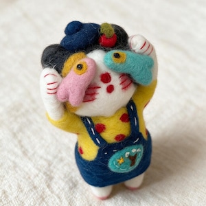 May include: A needle-felted cat figurine wearing a dark blue beret with a red cherry, a yellow polka-dot shirt, and dark blue overalls. The cat holds a small pink fish over its left eye and a light blue fish over its right eye, with red whiskers.