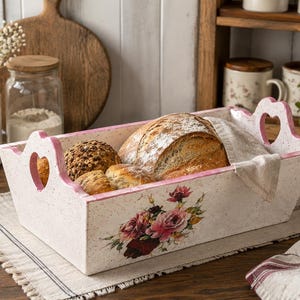 Puede incluir: Una cesta de pan rectangular de madera con acabado blanco desgastado, bordes pintados en rosa y recortes en forma de corazón en las asas. El frente presenta un diseño floral de rosas rosas y rojas. Contiene varios panes y un paño de color beige.