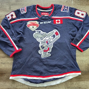 Peut inclure: Un maillot de hockey bleu marine CCM avec un graphique représentant un personnage de dessin animé portant un chapeau à larges bords et tenant une crosse de hockey. Le maillot porte les inscriptions Scotiabank Hockey Club, Rebels et le numéro 8.