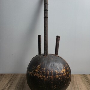 May include: A dark brown, spherical, hollow instrument with a long, vertical wooden neck and two shorter, parallel wooden pegs extending from the top. The surface of the sphere shows signs of wear with visible scratches and lighter brown patches.
