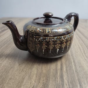 以下が含まれることがあります： 光沢のある仕上げと蓋が付いたダークブラウンの陶器製ティーポット。本体には、花柄や垂れ下がったタッセルなどの複雑な金色の模様が施されており、白、青、オレンジの小さな盛り上がったドットでアクセントが付けられています。