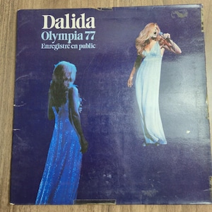 May include: A square vinyl record cover with a deep purple background featuring the text Dalida Olympia 77 Enregistré en public. The cover displays two images of a person with long hair in a floor-length gown, one singing into a microphone.