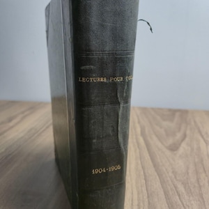 Könnte beinhalten: Ein dunkelgrünes gebundenes Buch mit strukturiertem Buchrücken. Der goldene Prägedruck auf dem Rücken lautet LECTURES POUR TOUS und 1904-1905. Das Buch weist Gebrauchsspuren mit sichtbaren Knicken am Rücken und ein kleines rot-schwarzes Lesezeichen auf.