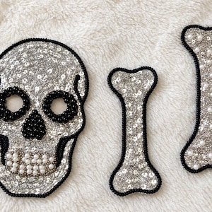 May include: A set of three embroidered patches featuring a skull and two bones. The skull has black beaded eye sockets and nose, with white pearl-like teeth. All pieces are covered in silver sequins and outlined with black beads.