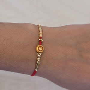 May include: A red thread bracelet featuring a central gold-toned circular charm with a swastika symbol, flanked by small gold-toned beads and elongated metallic accents. The bracelet is tied around a wrist, with a decorative card visible in the background.