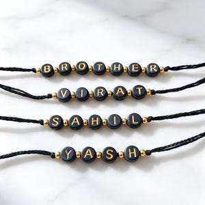 May include: Four black cord bracelets featuring round black beads with gold lettering spelling BROTHER, VIRAT, SAHIL, and YASH. Each bracelet is accented with small, round, metallic gold spacer beads between the lettered beads on the black string.