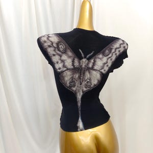 May include: A black short-sleeved top with a textured, pleated fabric surface. The back features a large, detailed graphic print of a moth in shades of white, gray, and black, with wings extending across the shoulders and a long tail design.