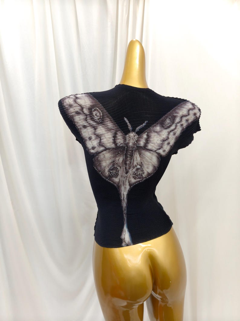May include: A black short-sleeved top with a textured, pleated fabric surface. The back features a large, detailed graphic print of a moth in shades of white, gray, and black, with wings extending across the shoulders and a long tail design.