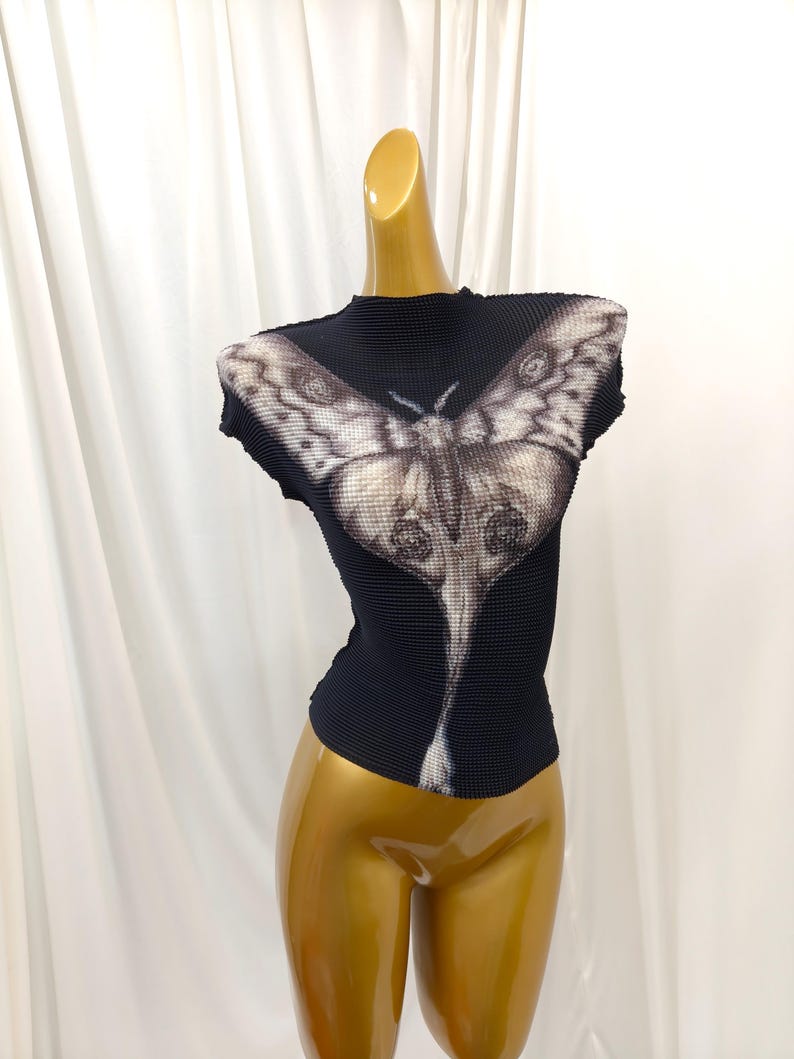 May include: A black, textured, short-sleeved top with a high neckline. The garment features a large, detailed graphic print of a moth in shades of grey, white, and black, extending from the chest down to the hemline of the fitted, pleated fabric.
