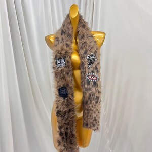 May include: A brown and black leopard print faux fur scarf featuring several decorative patches. The patches include a black square with white Japanese text and the word GRASS, a black cross, a red eye, and a black fabric rose flower.