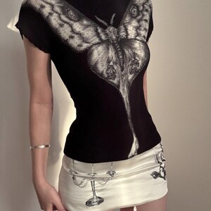May include: A black short-sleeved top with a high neck and a large white moth graphic across the chest, paired with a cream-colored mini skirt featuring a black and white illustration of a candelabra and other decorative elements on the fabric.